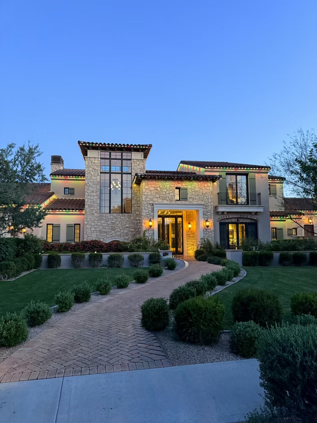 Modern stone-and-wood house with lit windows, landscaped front yard, and curved driveway at dusk