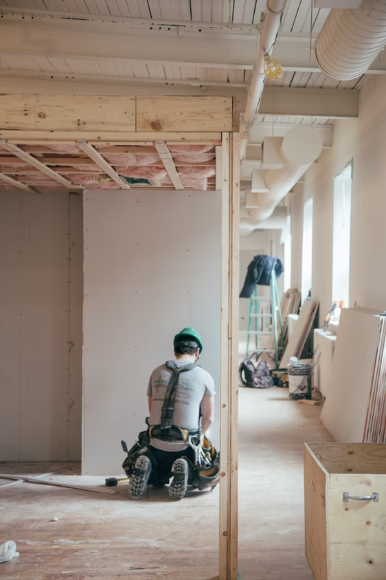 Worker kneeling in a partially finished room with exposed framing and drywall during interior renovation