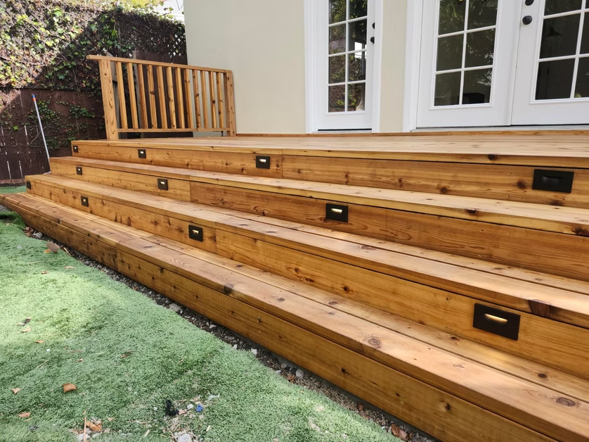 Wooden multi-level deck steps with built-in lights beside a white house and grass lawn