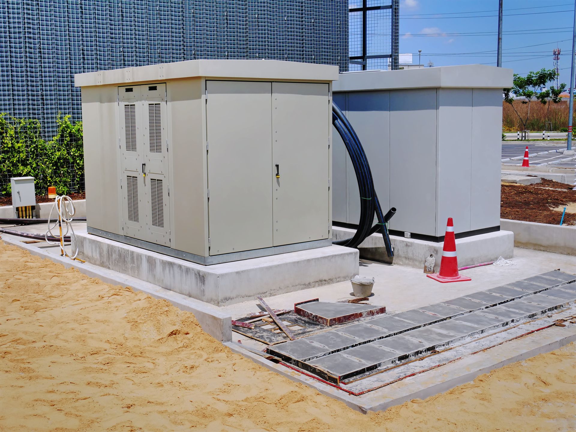 Construction site with two white utility boxes, cables, traffic cones, and paving stones on sandy ground