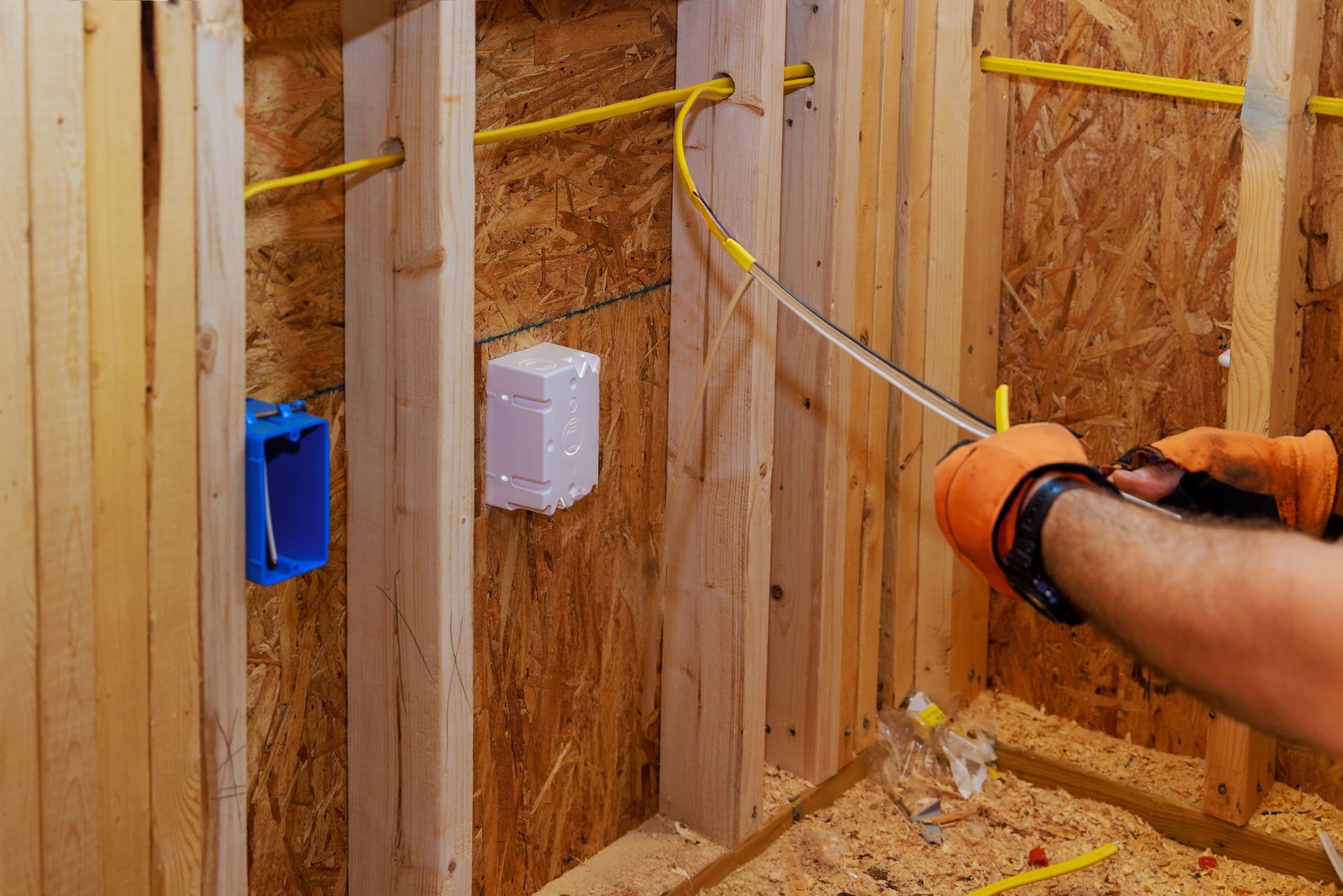 Person stripping electrical wire inside a wood-framed wall with blue and white outlet boxes.