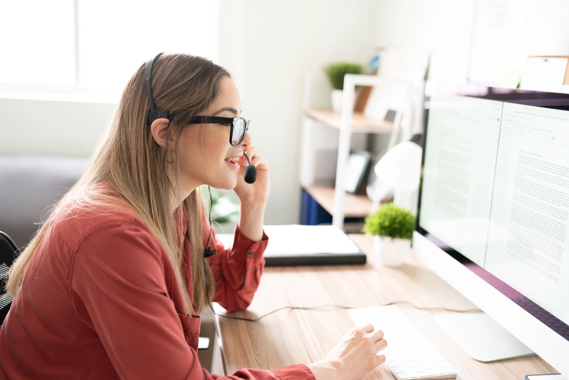 Woman wearing glasses works at a computer in a bright office, holding a phone to her ear.