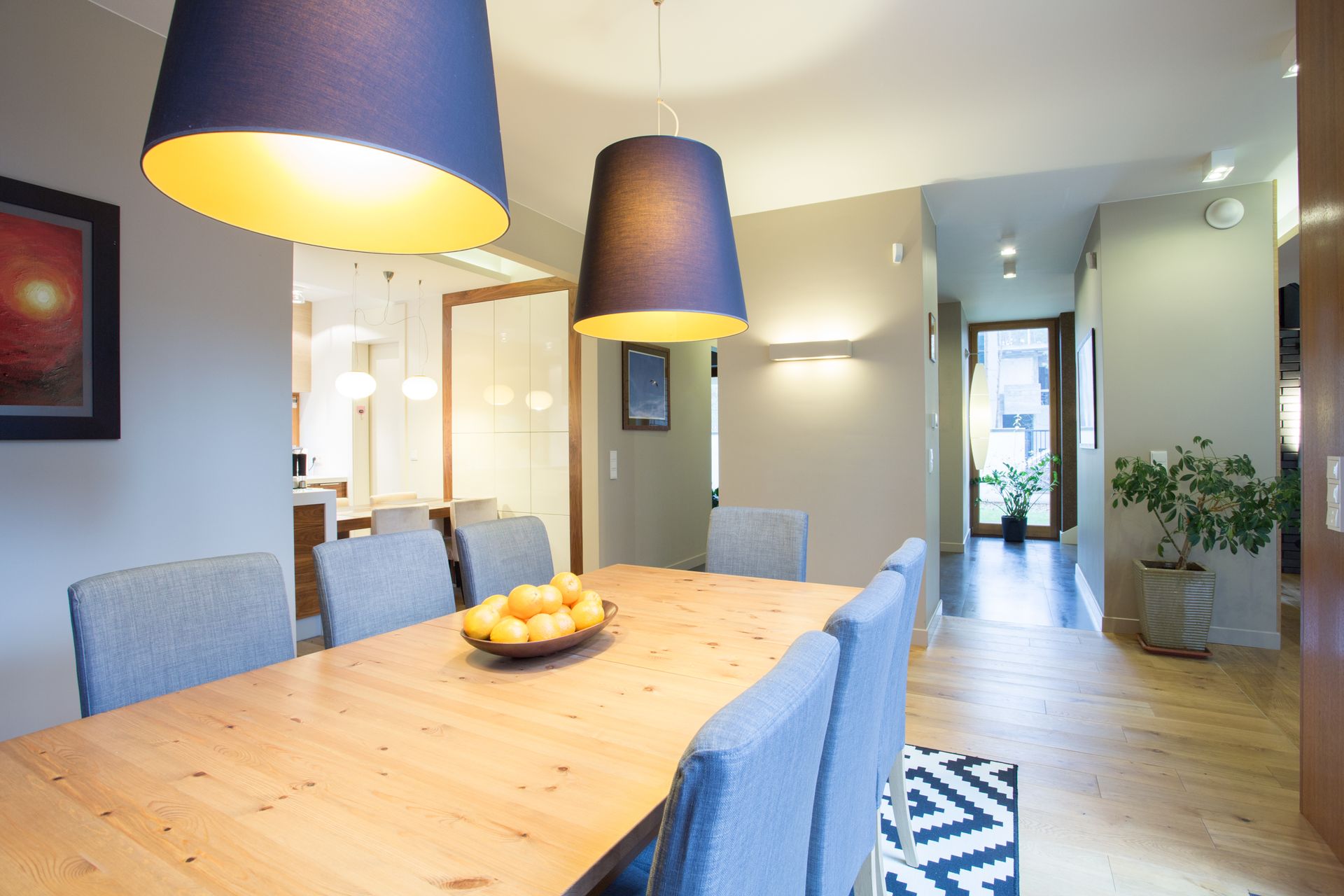 Bright dining room with wooden table, blue chairs, hanging lamps, and a bowl of oranges