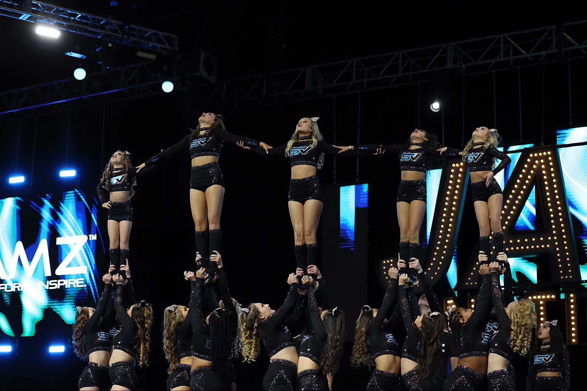 Cheerleaders in black uniforms perform a pyramid. Stage lit with bright lights and a large letter 