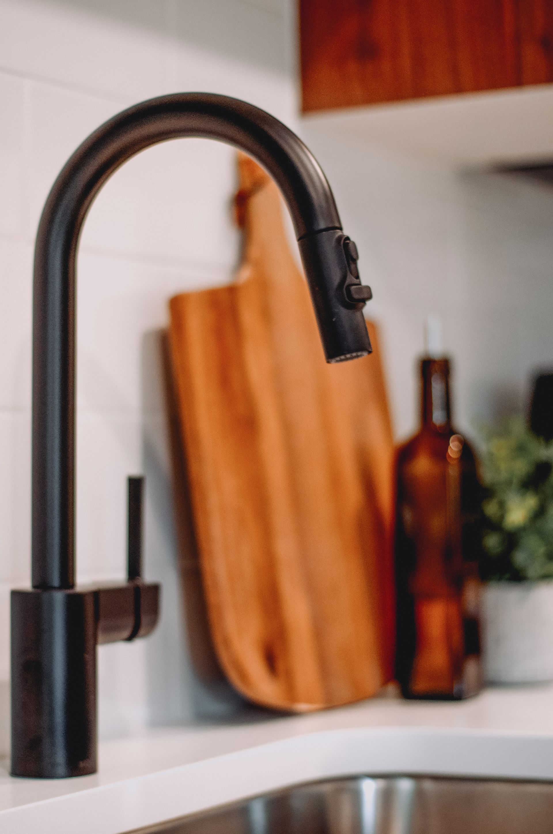 Black kitchen faucet with a pull-down sprayer. Cutting board, brown bottle, and plant sit behind it.