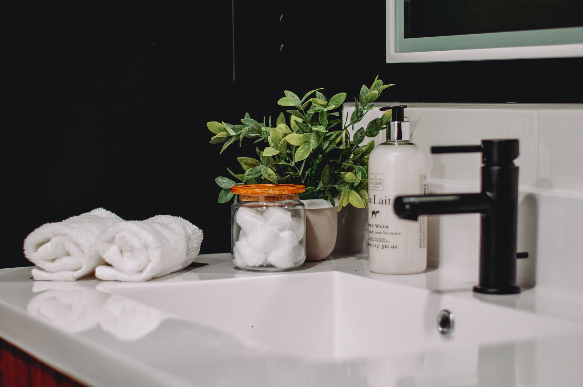 Bathroom sink with rolled white towels, green plant, jar of cotton balls, soap, and black faucet.