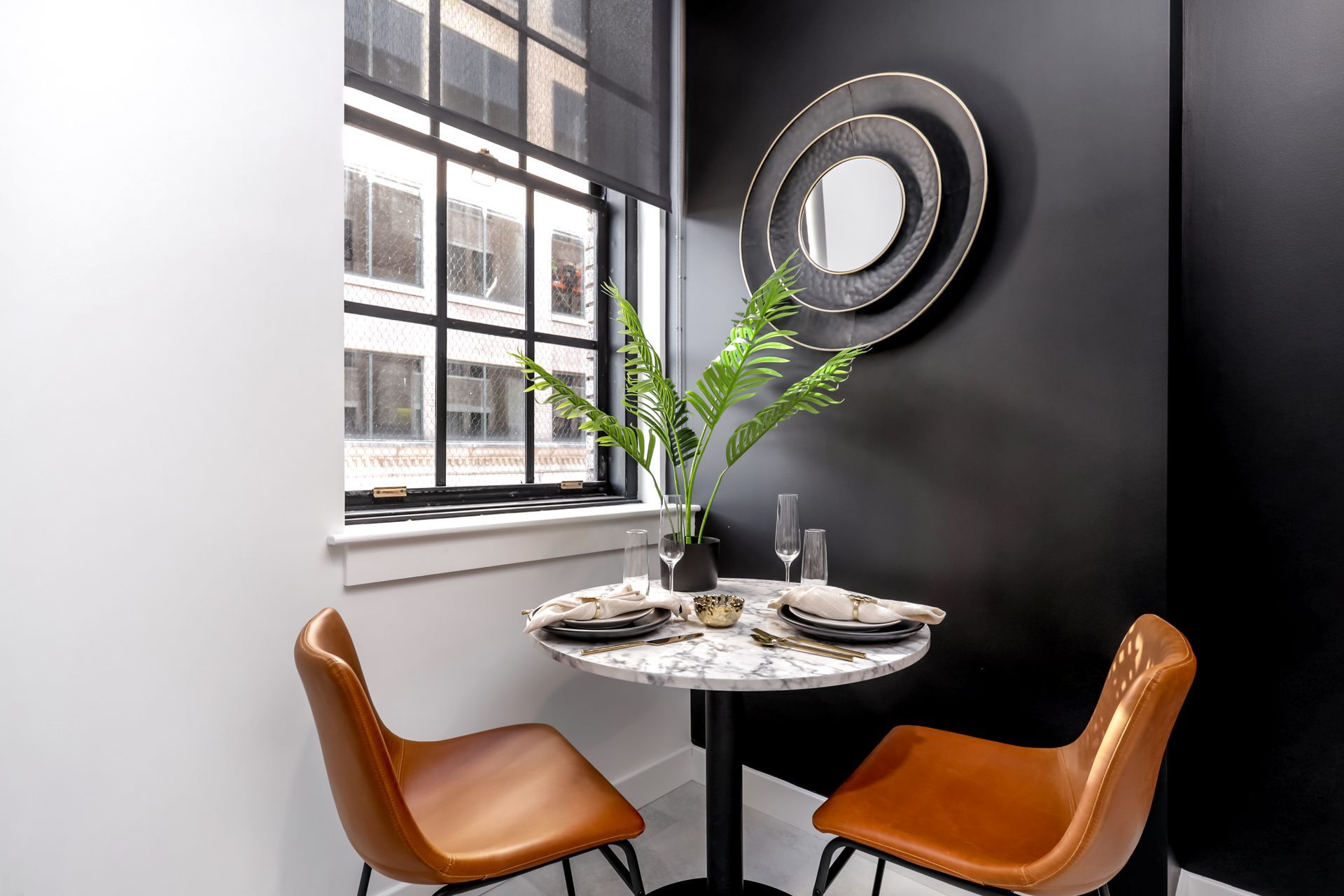 Small dining nook with marble table, two tan chairs, and a black-framed window. Black wall with a decorative mirror.