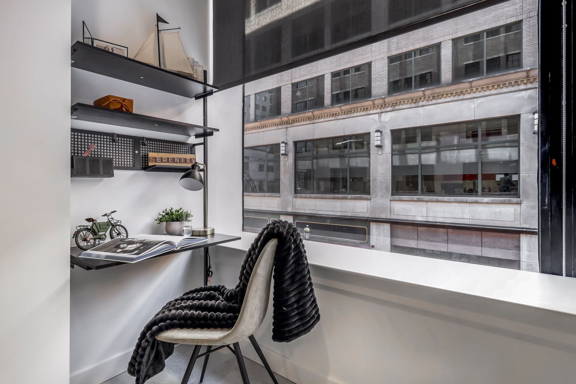 Desk nook with city view, shelves with decor, and a black blanket draped over a chair.