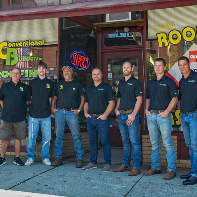 Six men in black shirts and jeans stand outside a storefront with an OPEN sign.