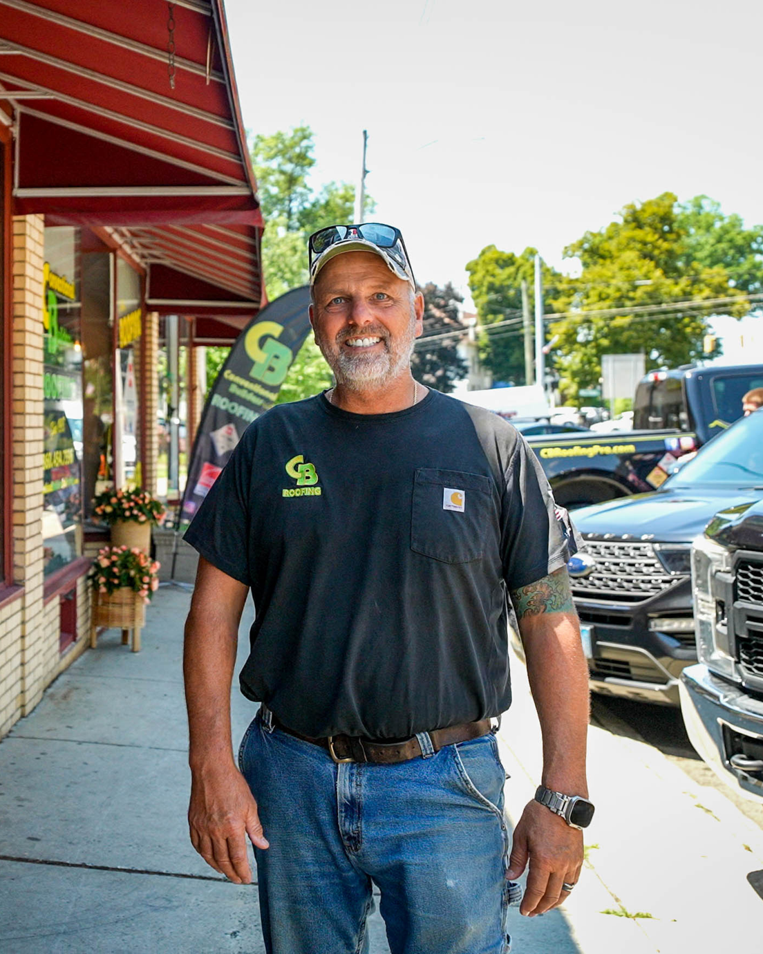 Man smiling on a sidewalk outside shops, wearing a black T-shirt, jeans, and a cap.