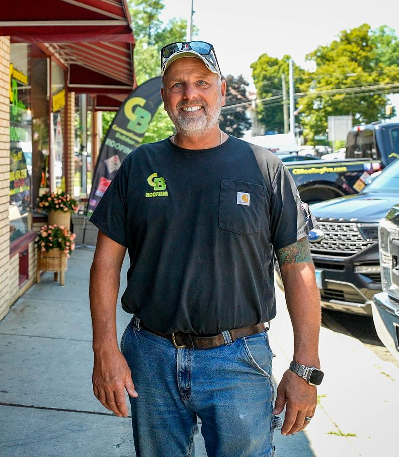 Man smiling on a sidewalk outside shops, wearing a black T-shirt, jeans, and a cap.