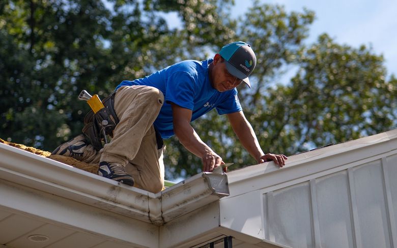 Worker on a roof repairing white gutters with tools, wearing blue shirt and cap.