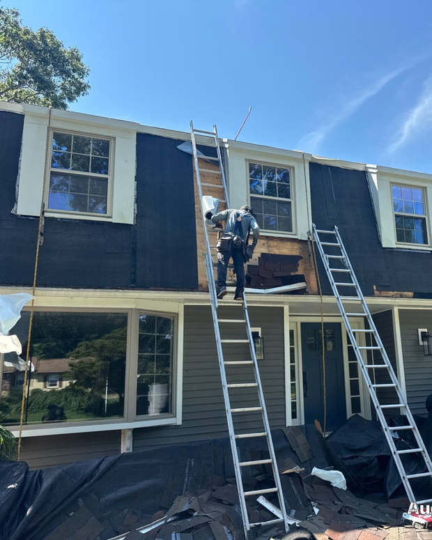 Workers on ladders repair the second-story exterior of a house with dark siding.