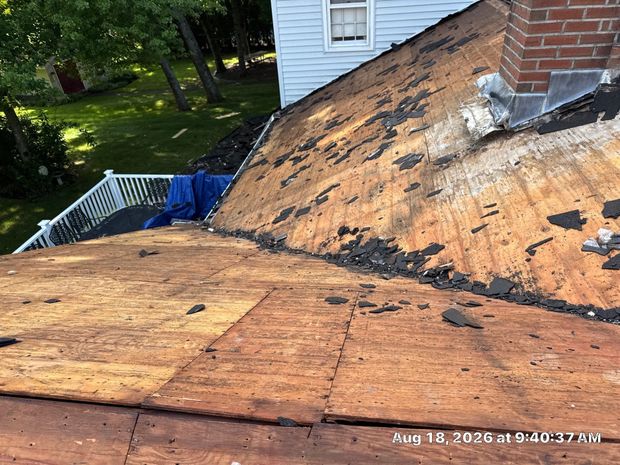 Partially stripped roof with exposed plywood and damaged shingles near a white house and chimney