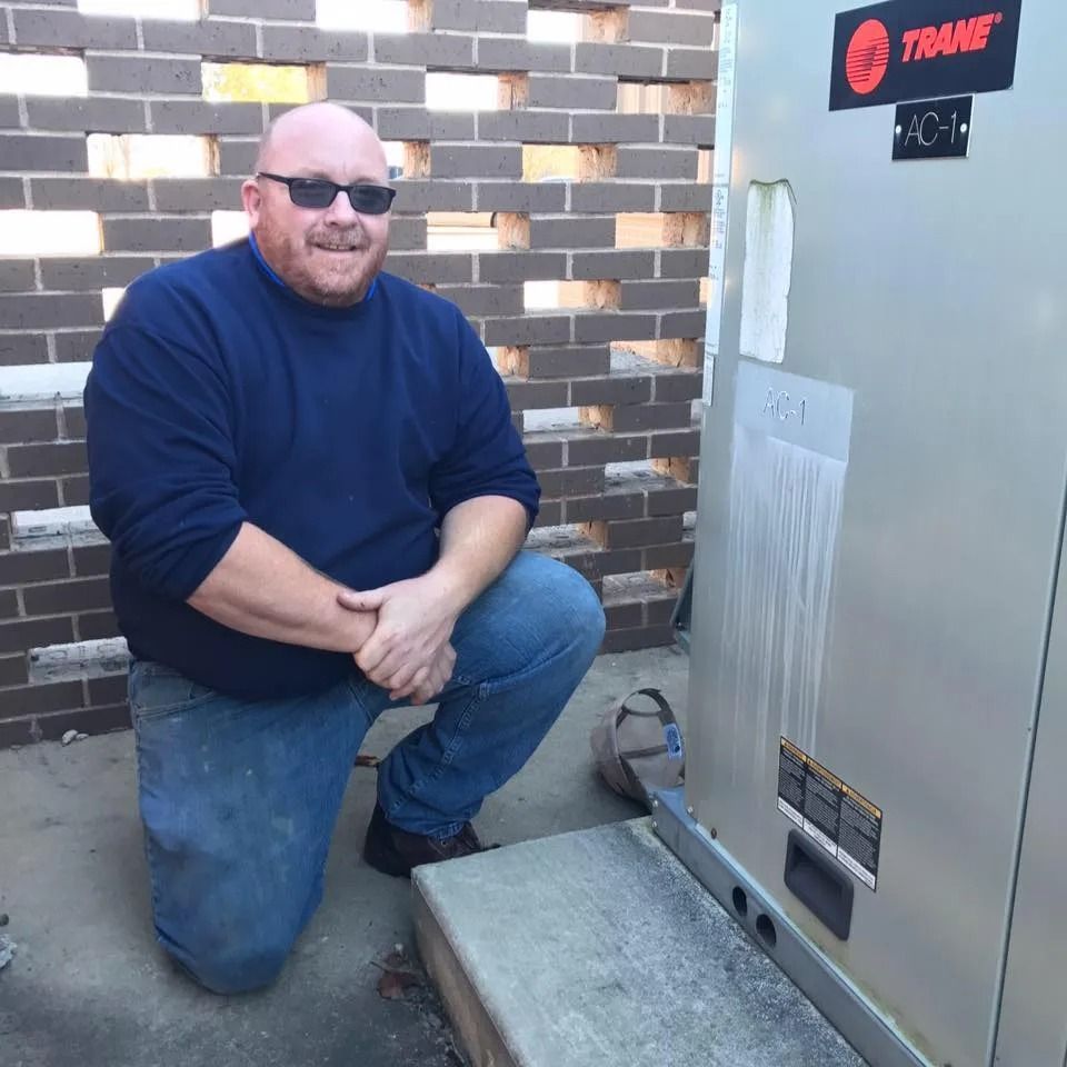 Man kneeling beside a Trane HVAC unit outdoors next to a brick wall and concrete slab.