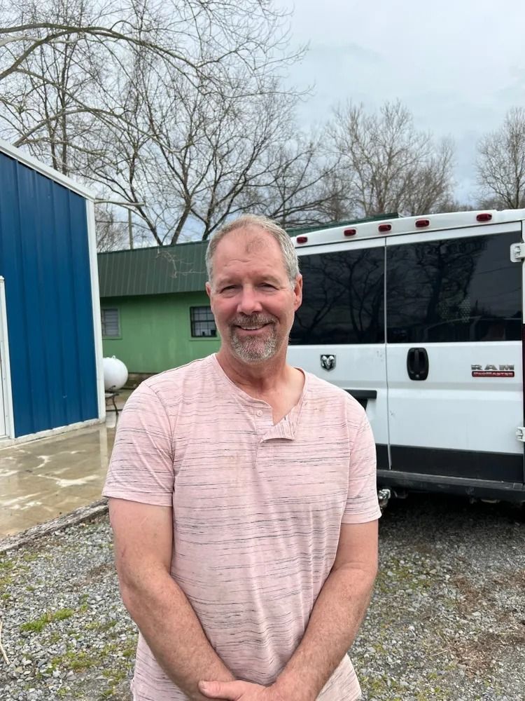 Man standing outdoors beside a white van and blue building on a cloudy day, smiling at the camera