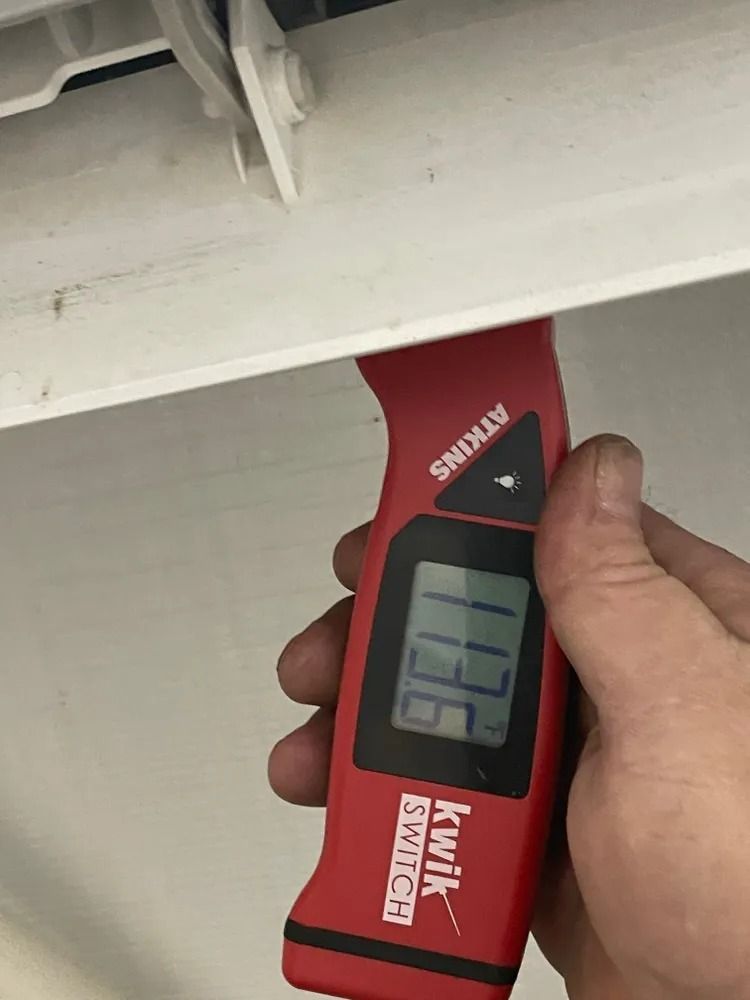 Hand holding a red digital moisture meter against a white surface under a beam