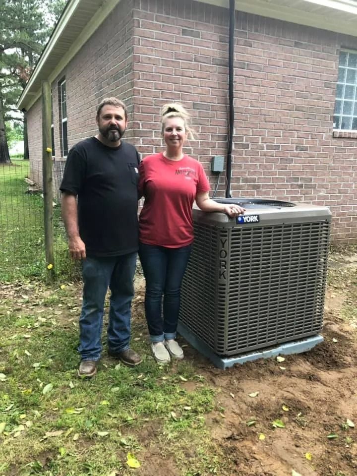 Two people standing beside a large outdoor air conditioner unit next to a brick house.