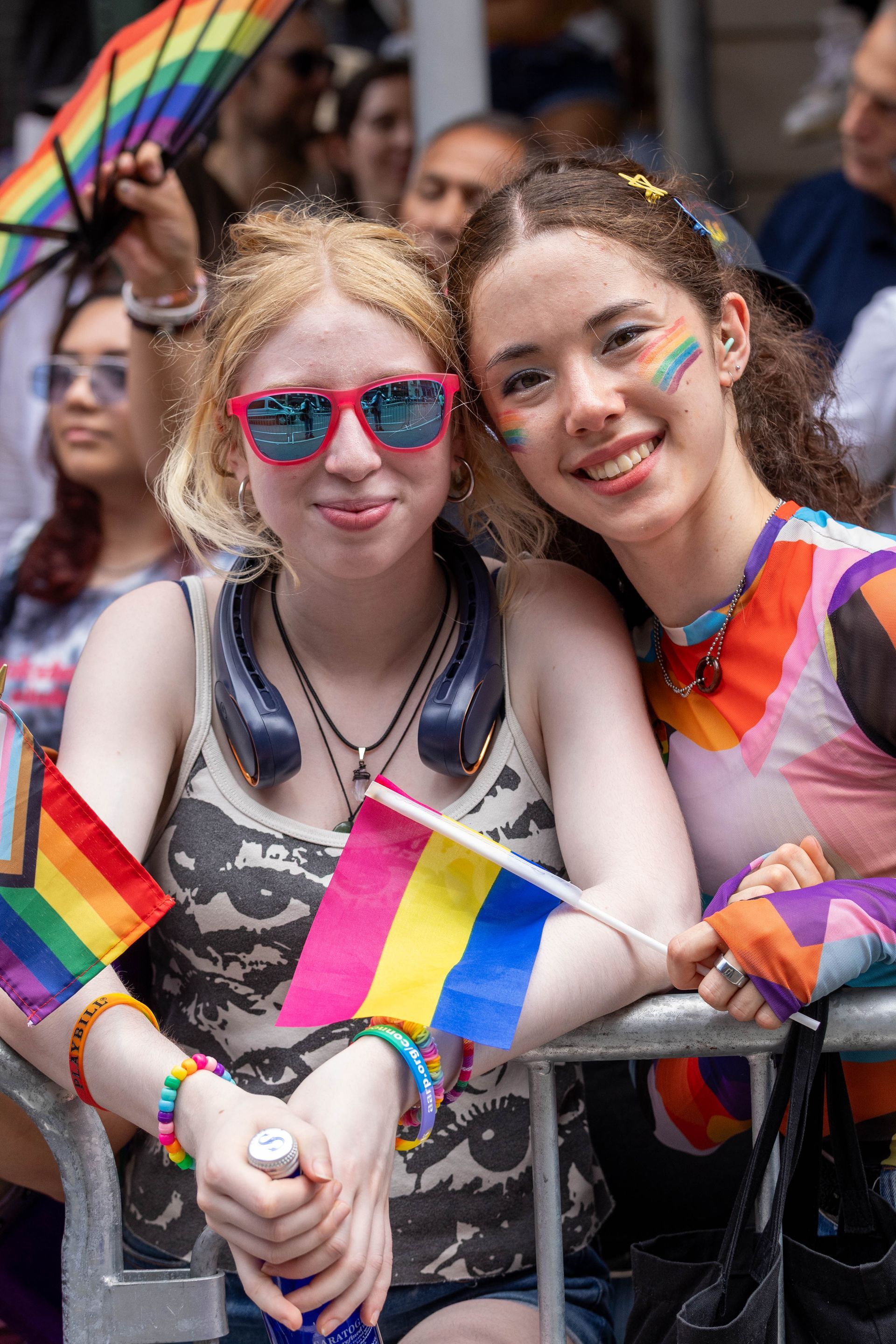 Two young people at Pride, holding flags. One wears rainbow makeup, the other sunglasses.