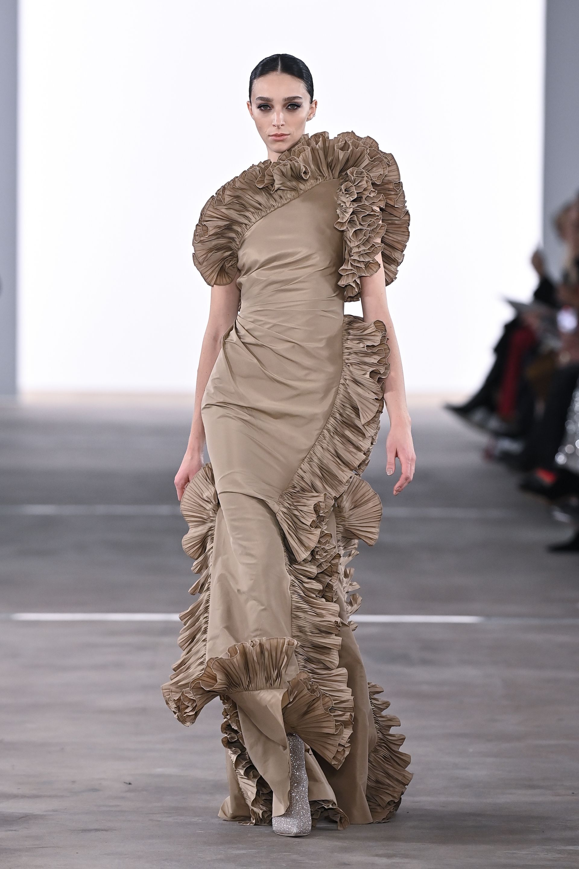 Model in a long, tan gown with large ruffles, walking a runway.
