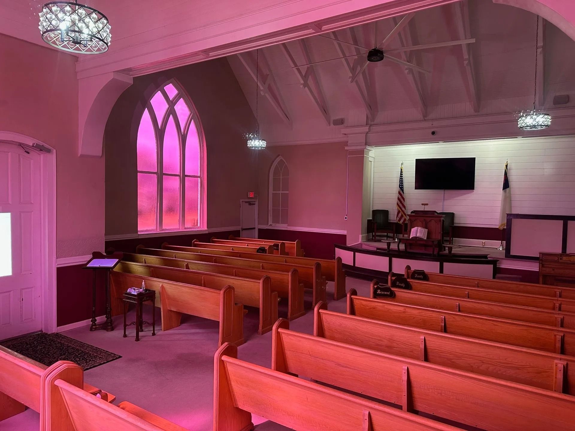 Pink-lit church sanctuary with pews, piano, pulpit, and stained-glass window