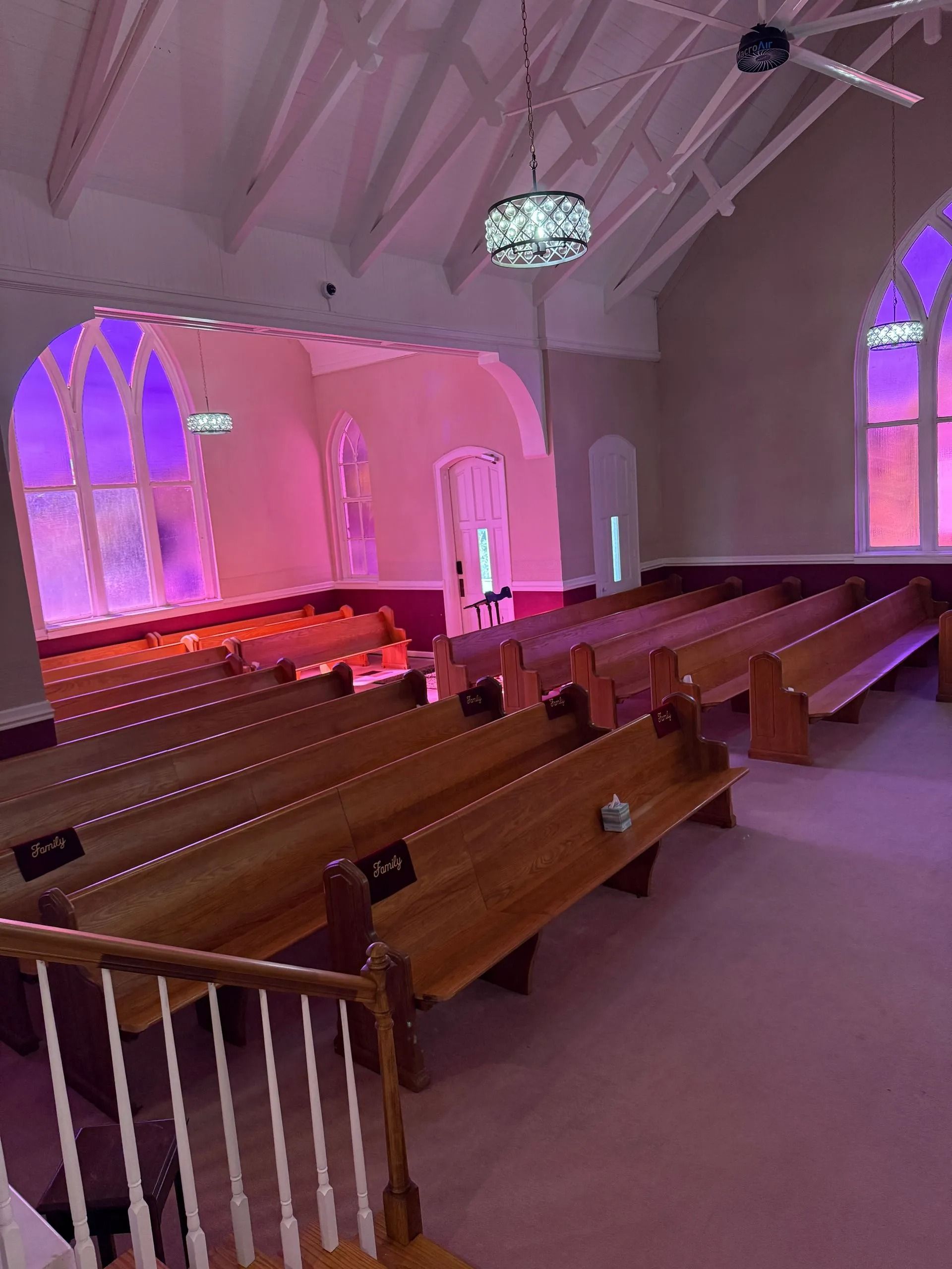 Empty church interior with wooden pews, arched windows, and pink-purple lighting