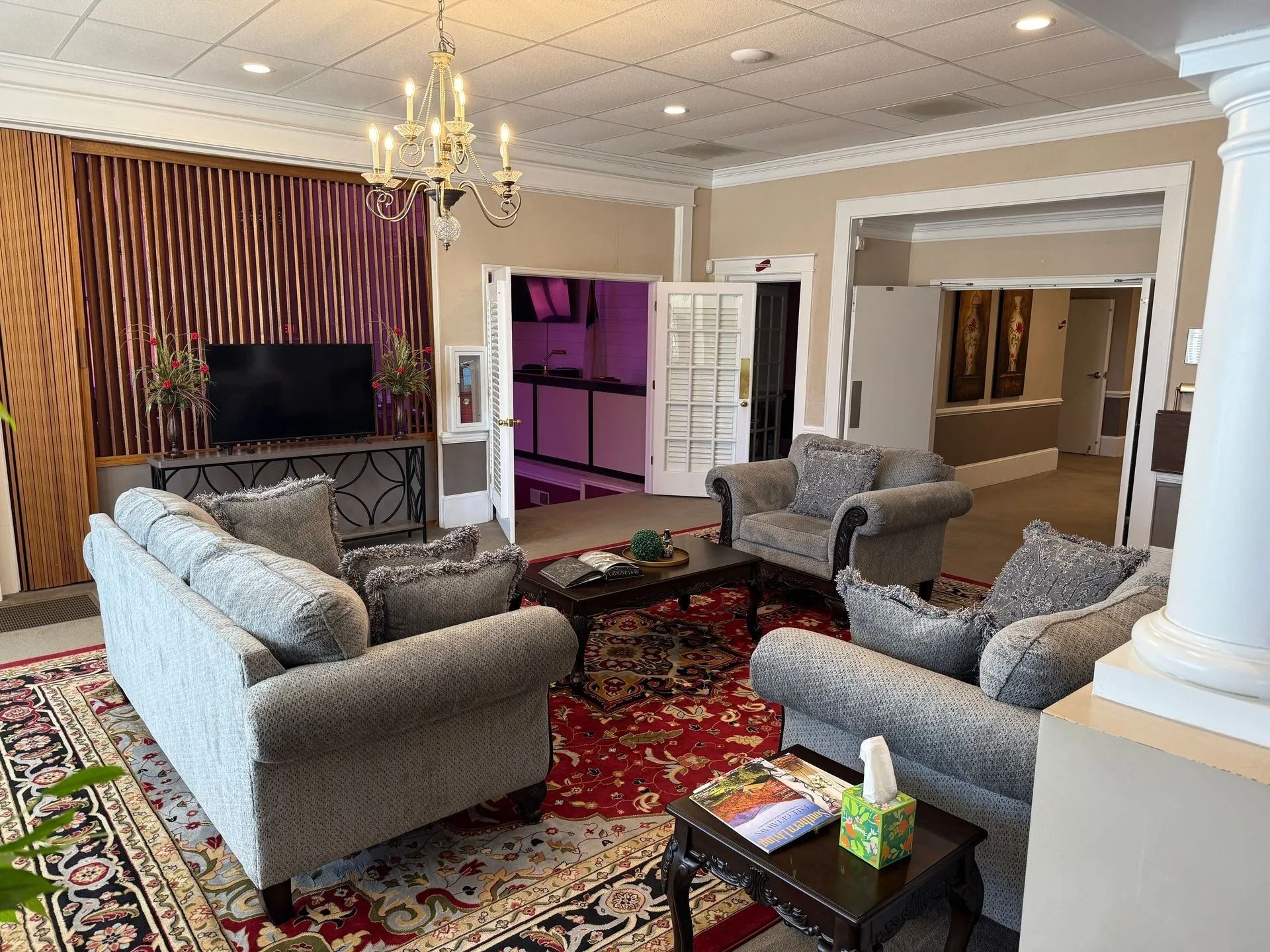 Living room with gray sofas, patterned rug, TV, chandelier, and purple-lit open doorway