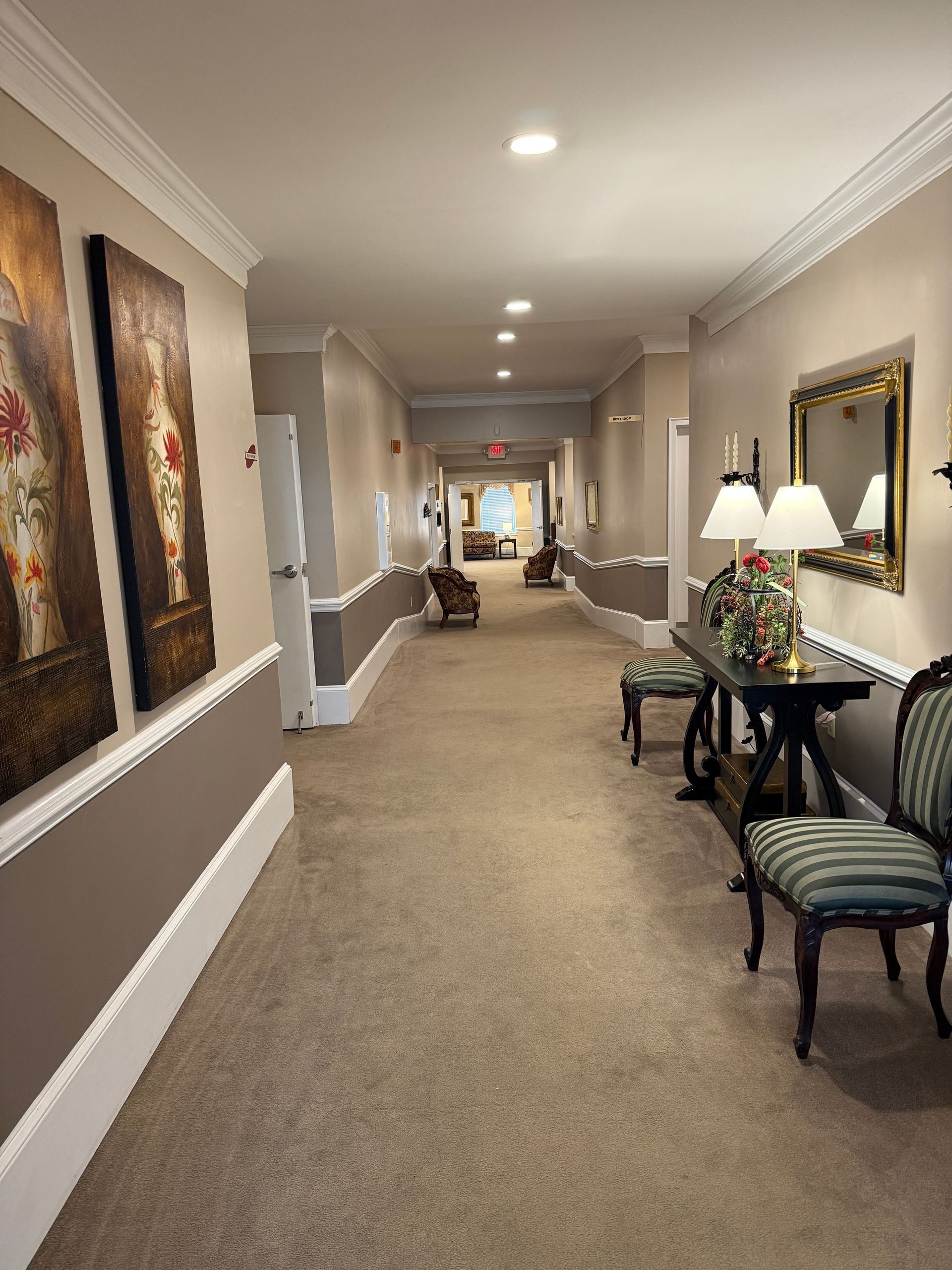 Carpeted hotel hallway with framed art, tables, lamps, and white wainscoting.