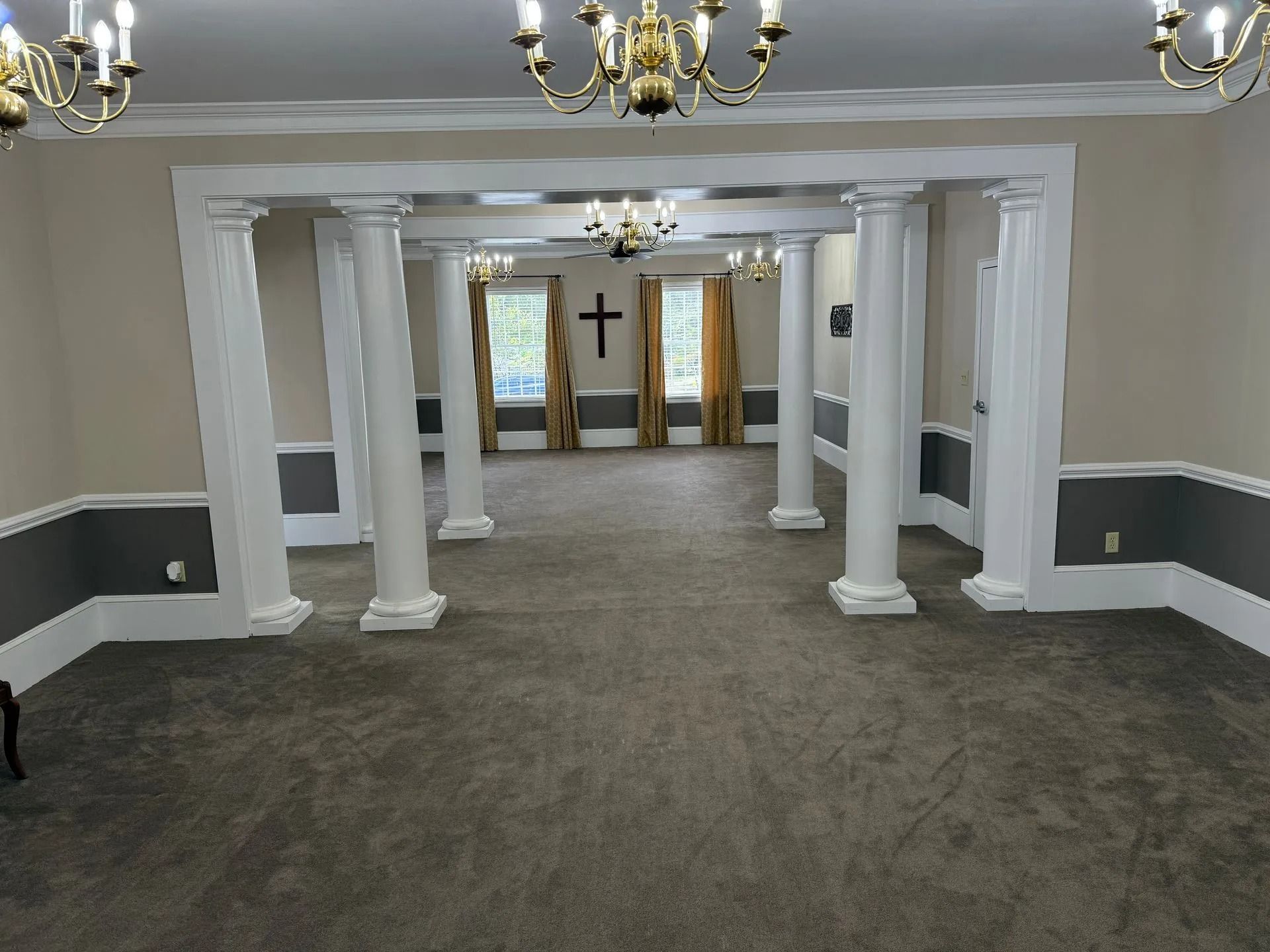 Empty church hall with white columns, chandeliers, carpeted floor, and a cross at the far end