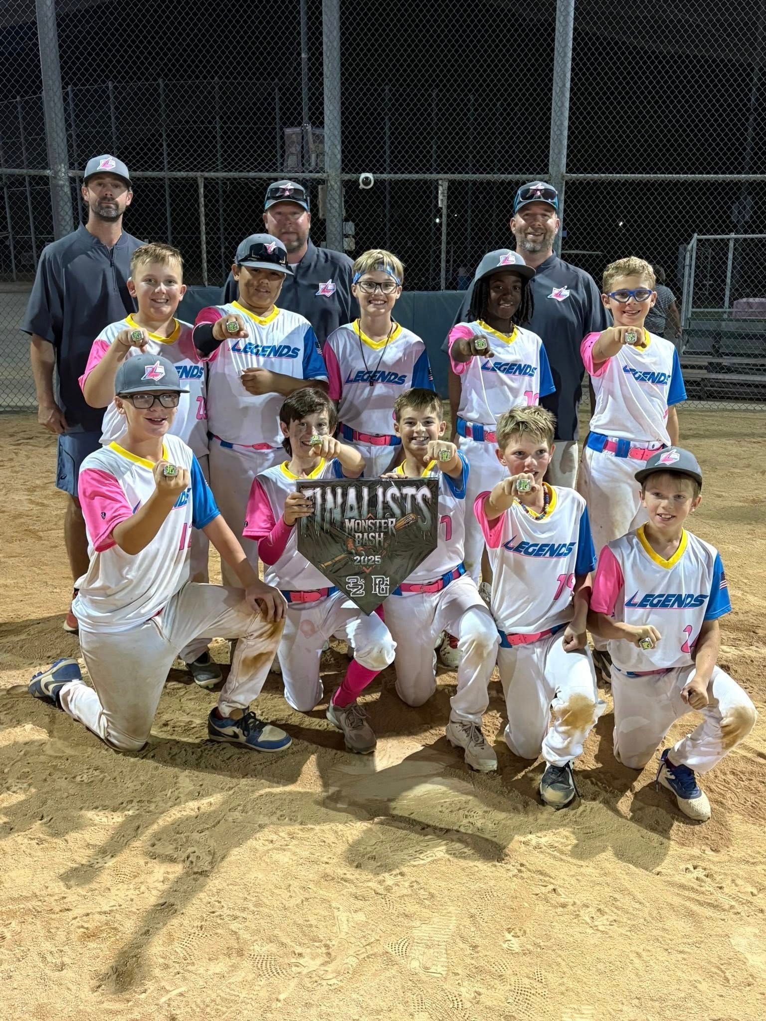 Youth baseball team posing with coaches on a dirt field, holding a championship banner.