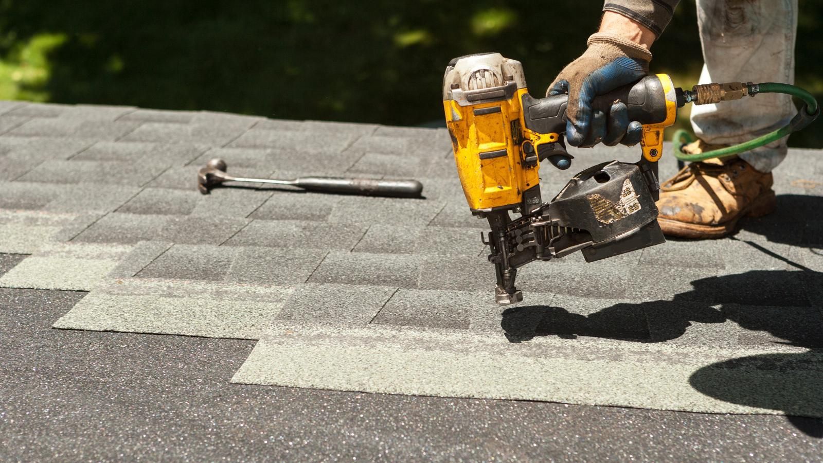 Worker using a yellow nail gun to fasten gray shingles on a roof