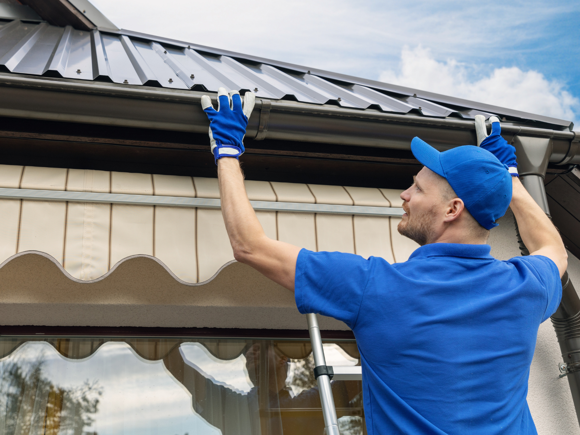 A man is standing on a ladder fixing a gutter on the roof of a house.