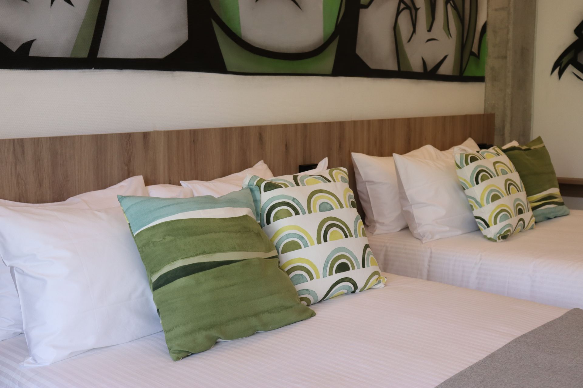 Two beds with white linens, decorative pillows in greens and blues, against a wood headboard, mural above.