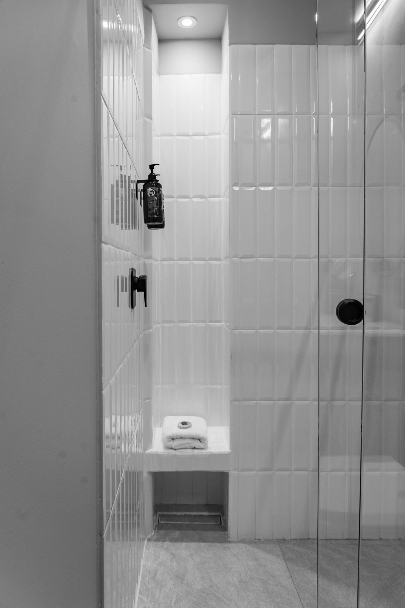 Black and white shower interior with white tiled walls, glass door, built-in shelving, and dispenser for soap.