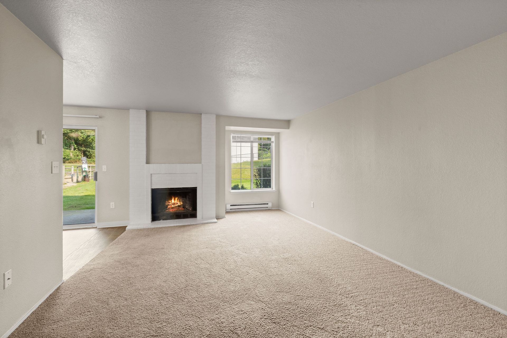 Empty living room with fireplace, sliding glass door, window, and beige carpet.
