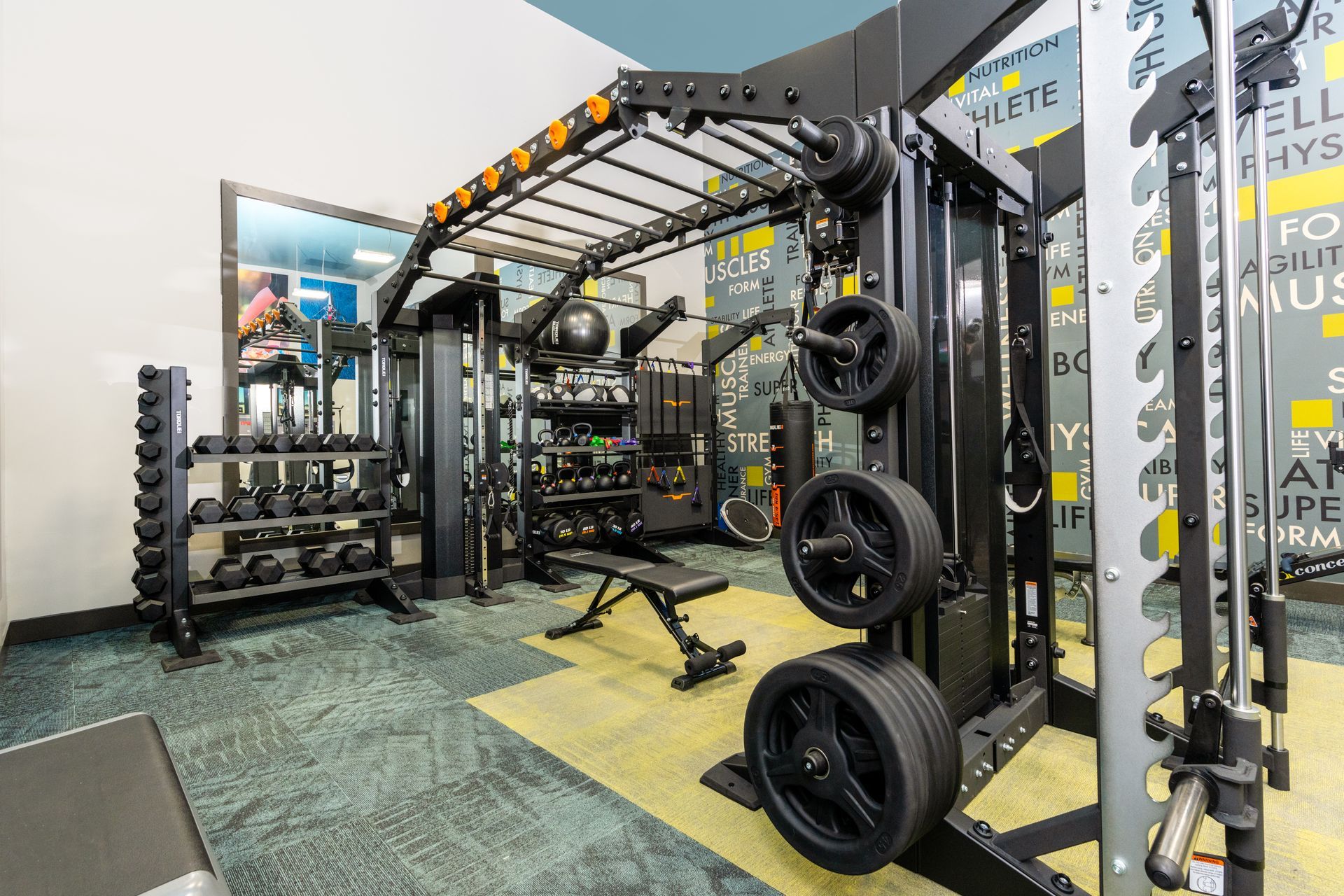 Gym interior with weight racks, bench, and equipment against a wall with inspirational text.