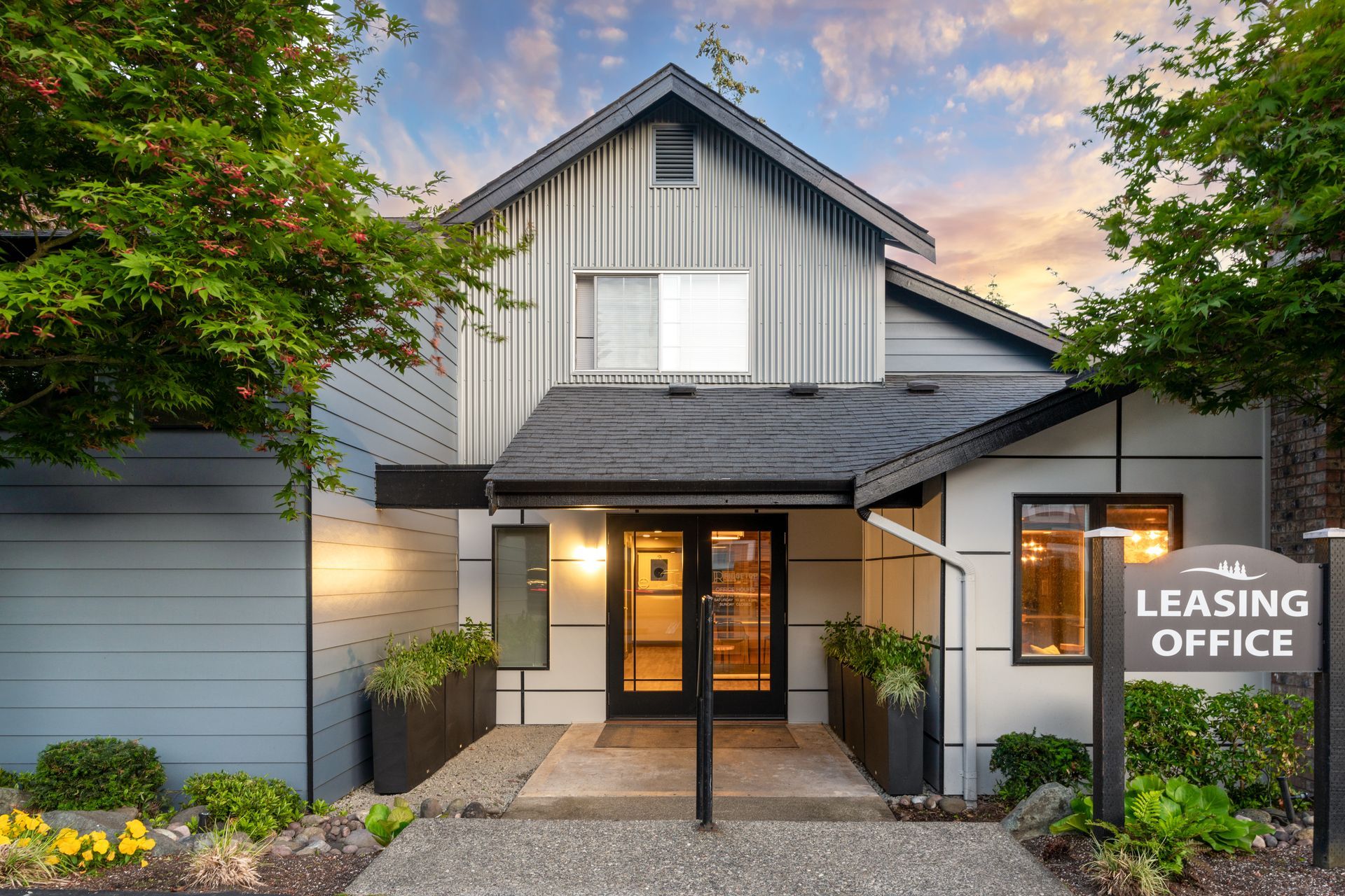 Leasing office entrance, gray and black building with sign, flanked by trees and greenery.