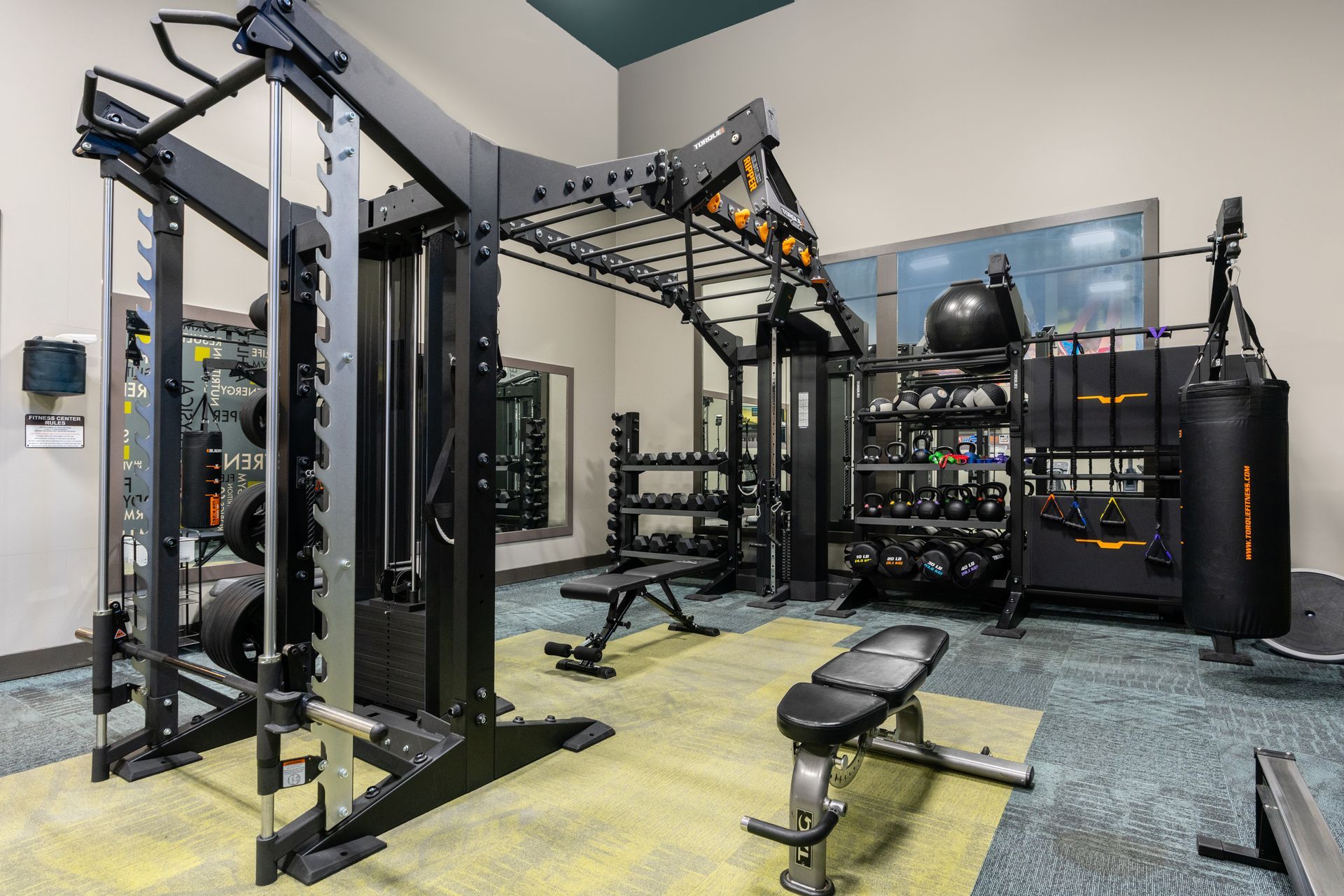 Gym with weight machines, free weights, benches, and a punching bag, on patterned carpet.