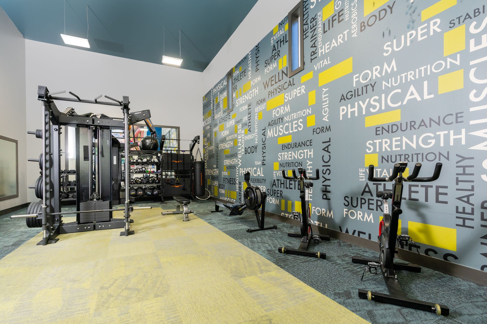 Gym interior with exercise machines: spin bikes, weights, and a multi-station weight machine, against a motivational wall.