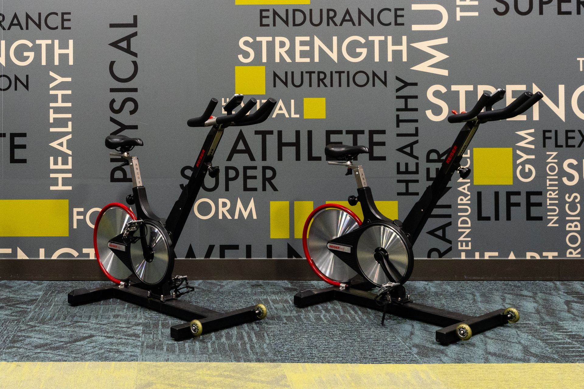 Two stationary bikes in a gym. Black frames, red flywheels, against a wall with fitness words.