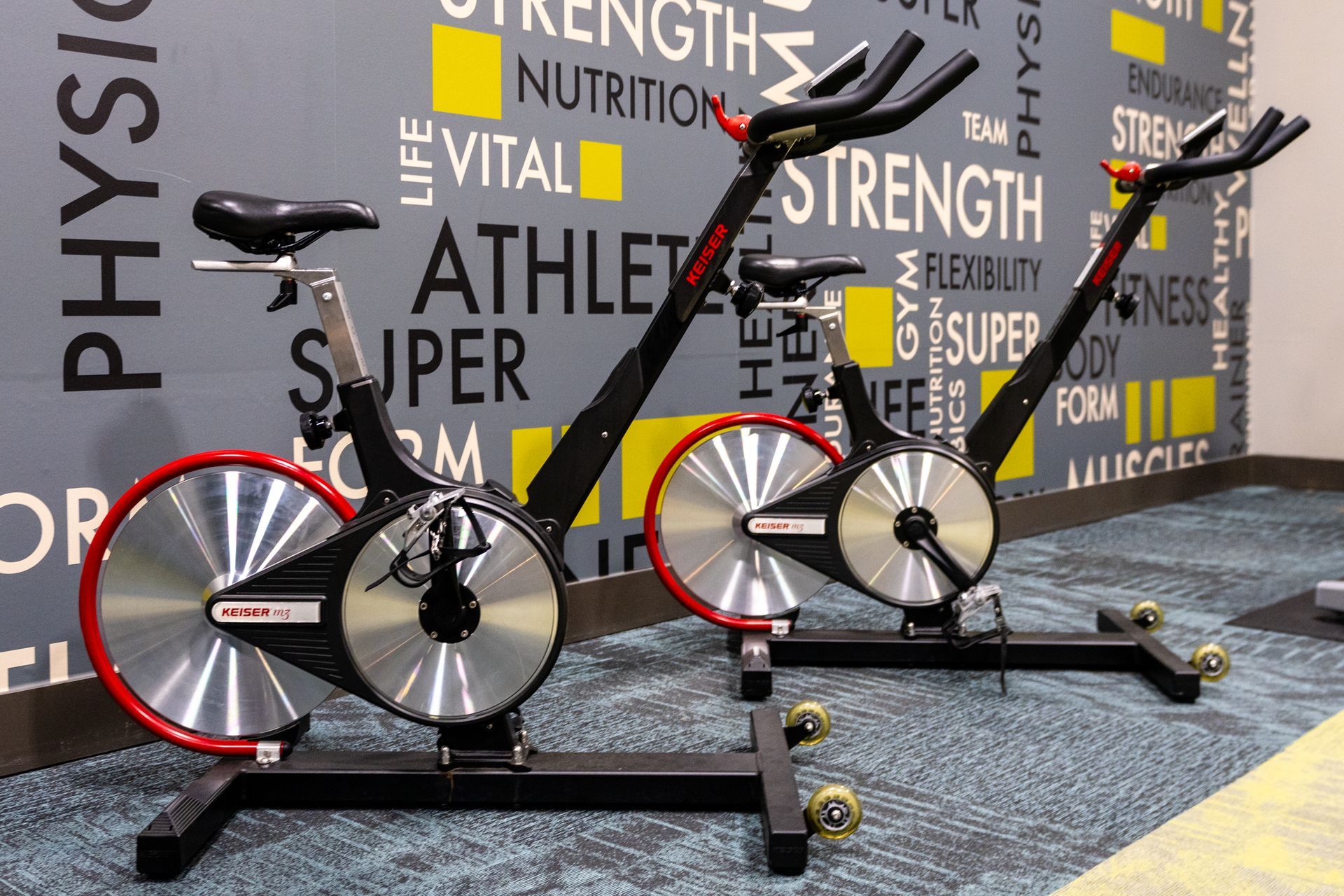 Two indoor cycling bikes with red and silver wheels sit in a gym with fitness-themed wall art.