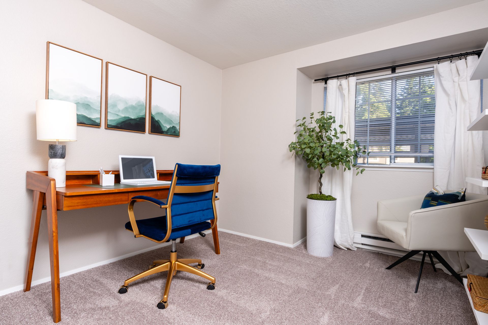 Home office with desk, laptop, blue chair, artwork, and window with curtains; light colors.