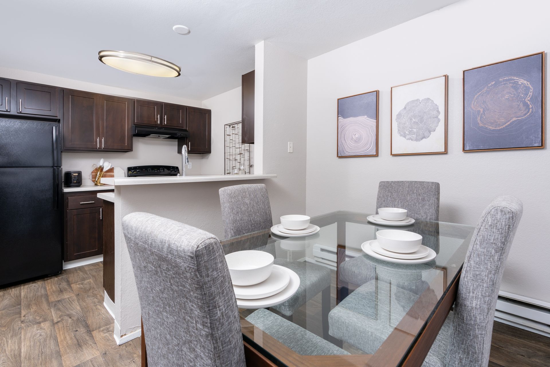 Dining area with glass table set for four; kitchen in background with dark cabinets and black appliances.