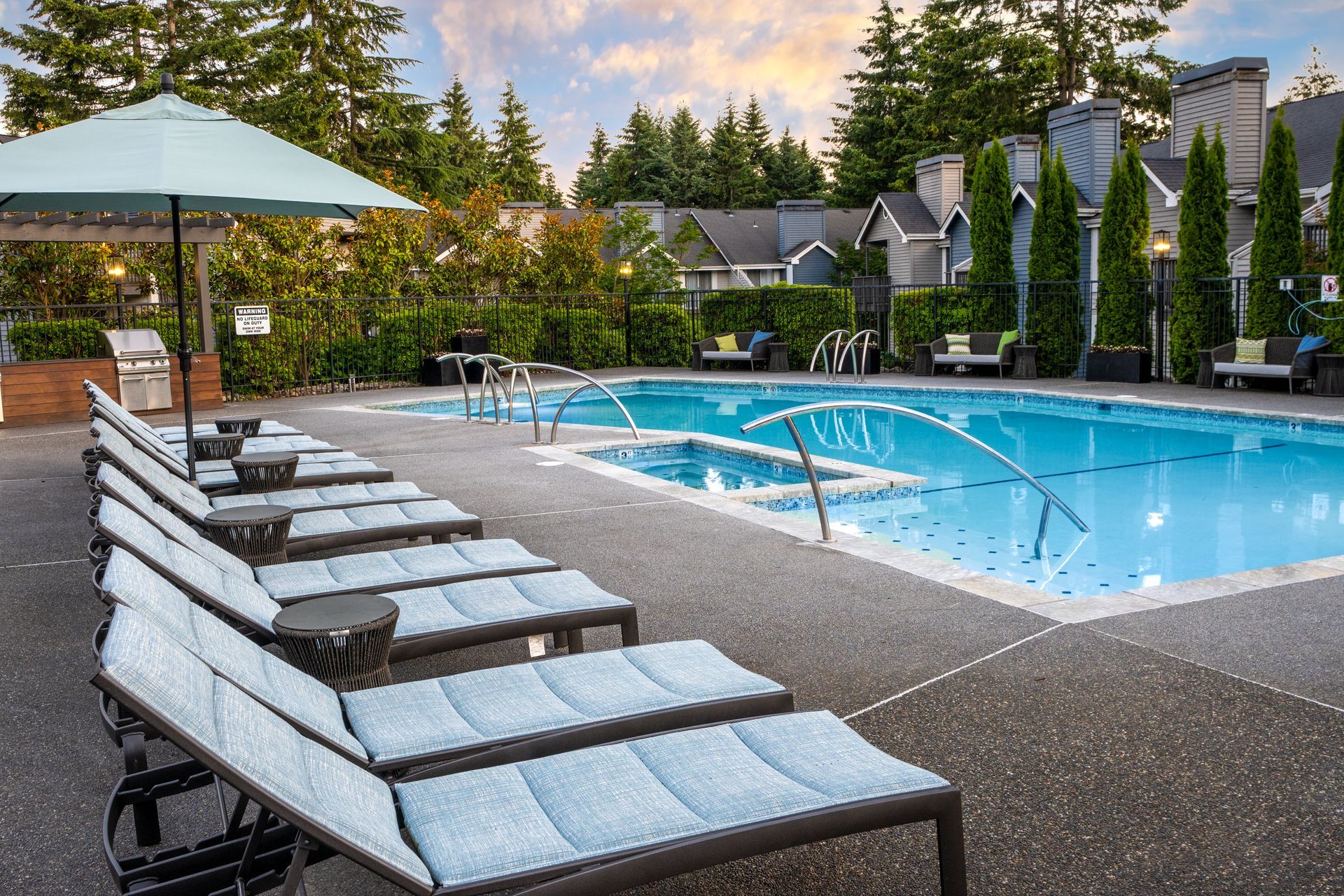 Poolside scene with lounge chairs, umbrella, and swimming pool. Houses and trees in background.