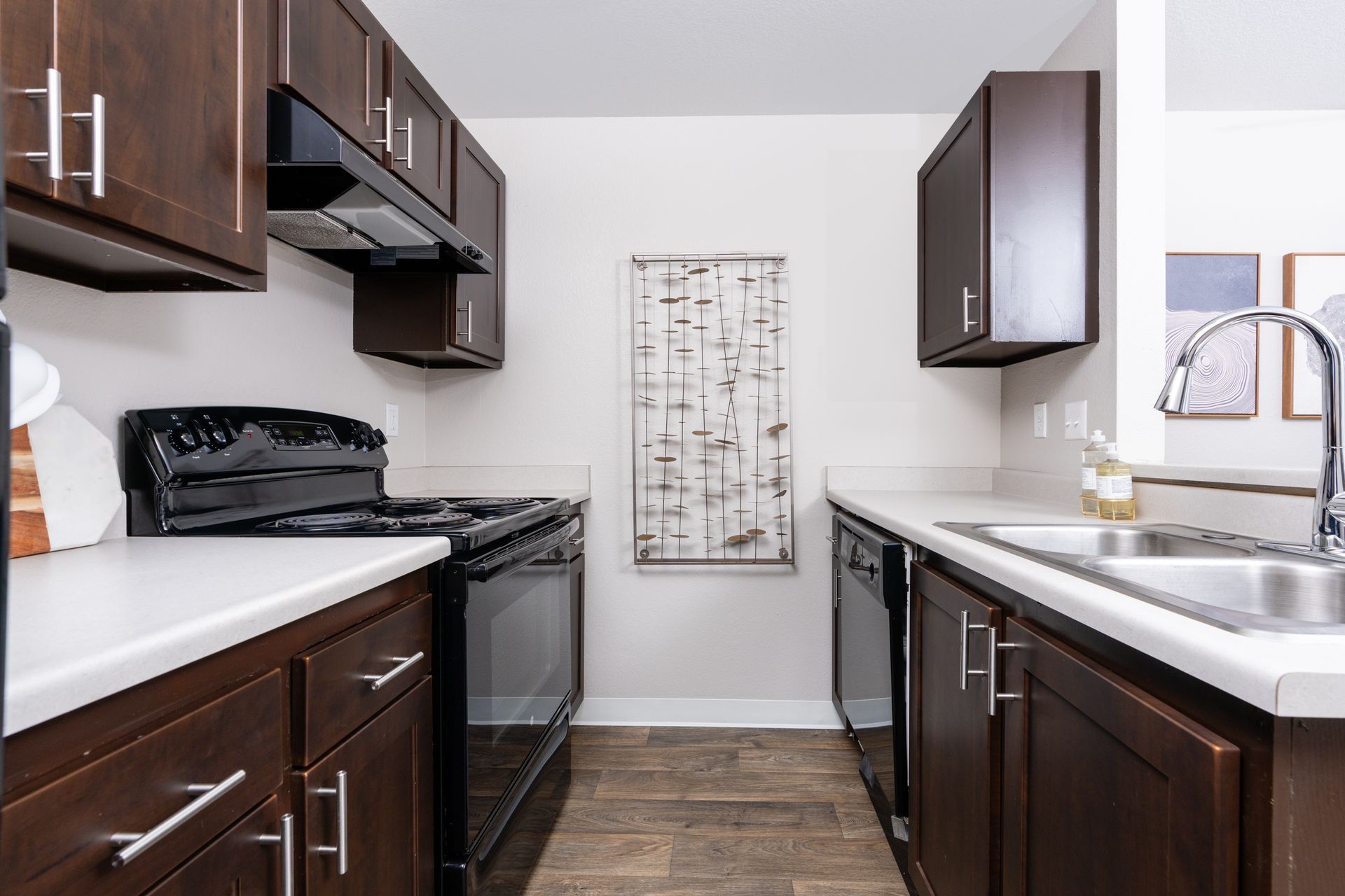 A narrow kitchen with brown cabinets, white countertops, black appliances, and a decorative piece on the wall.
