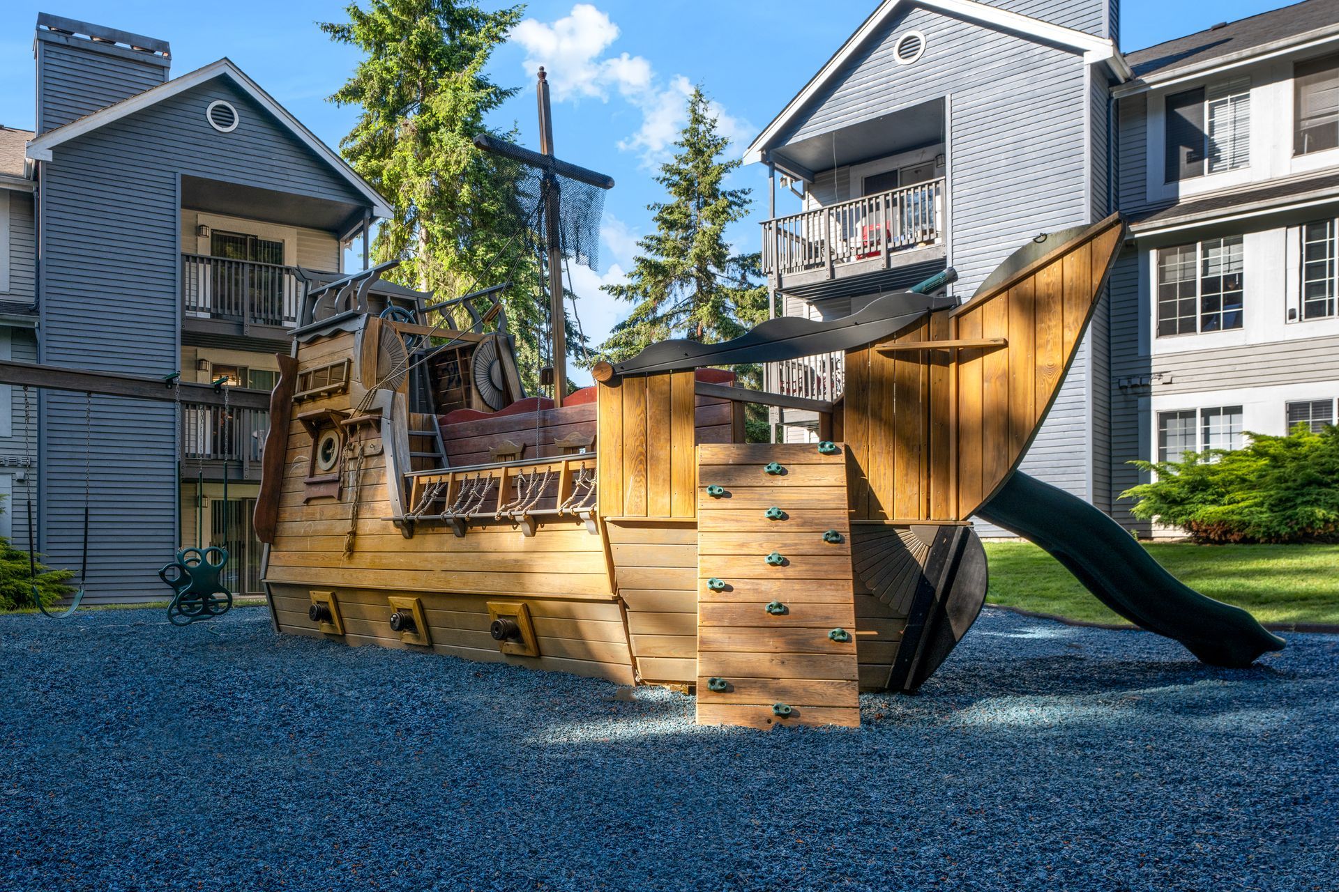 Playground pirate ship with slide and climbing holds in front of apartment buildings.