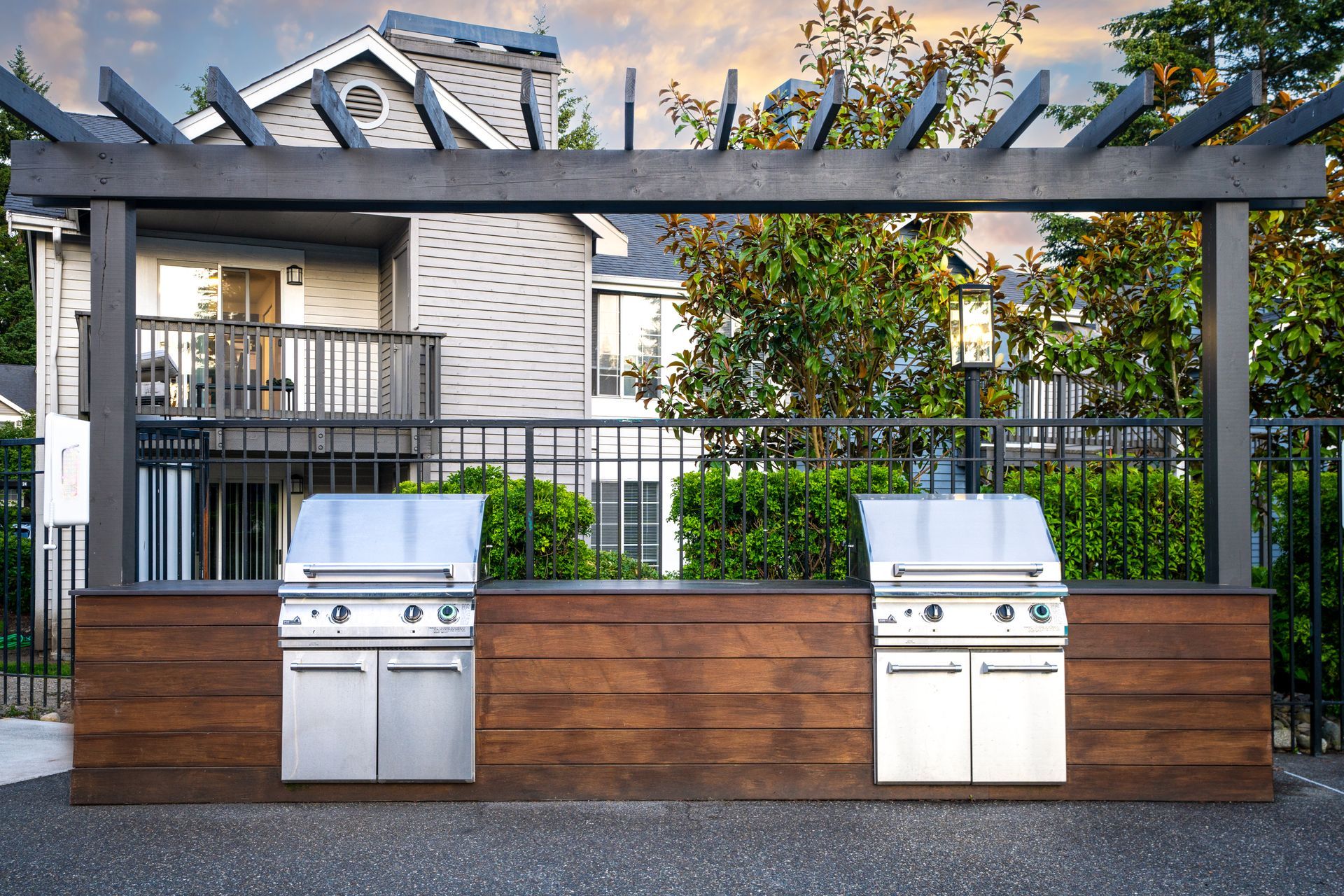 Outdoor grilling station with two stainless steel grills, brown wooden counter and pergola in front of apartment building.