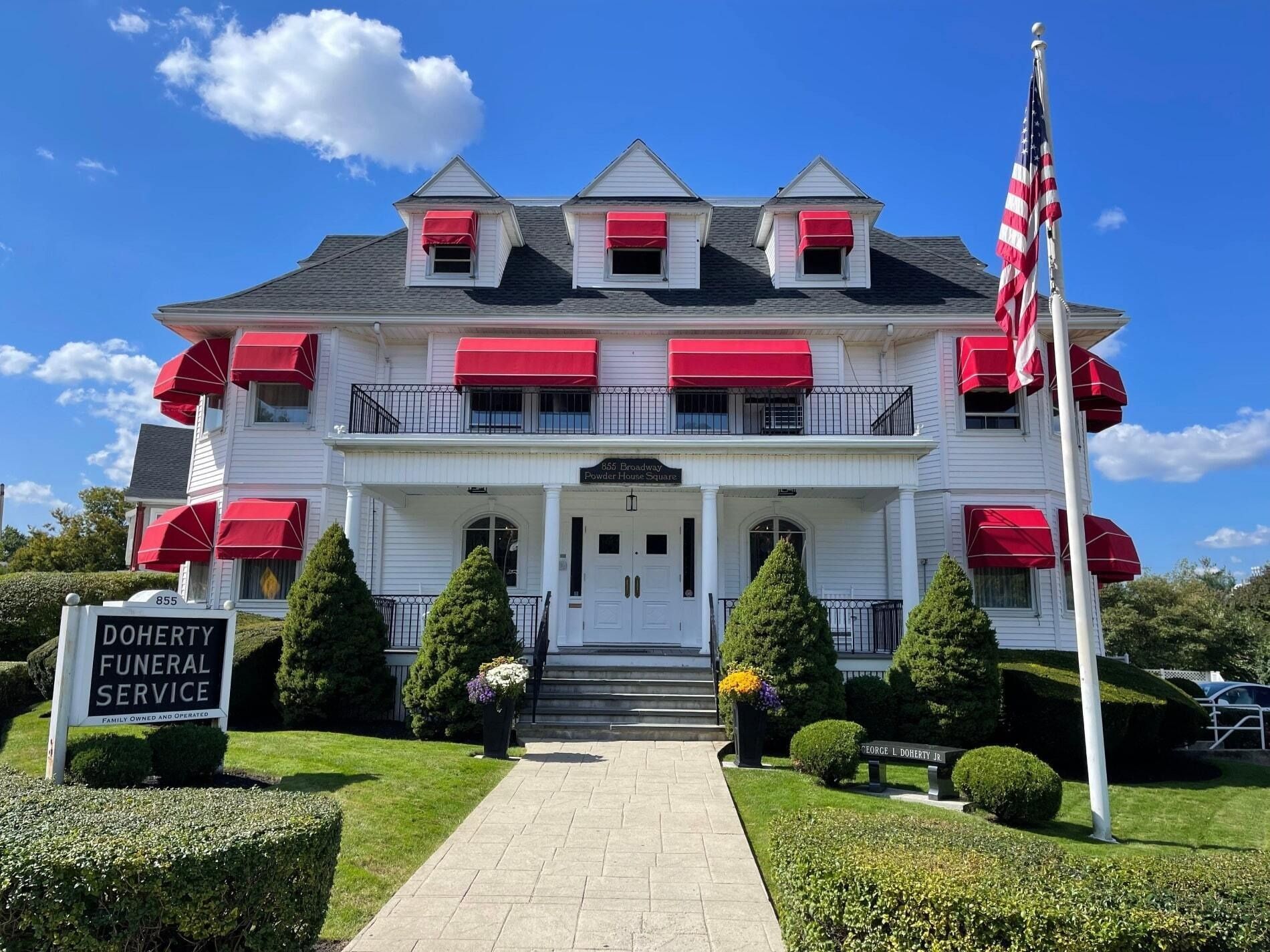 A white funeral home with red awnings and an American flag, viewed from a front walkway under a blue sky.