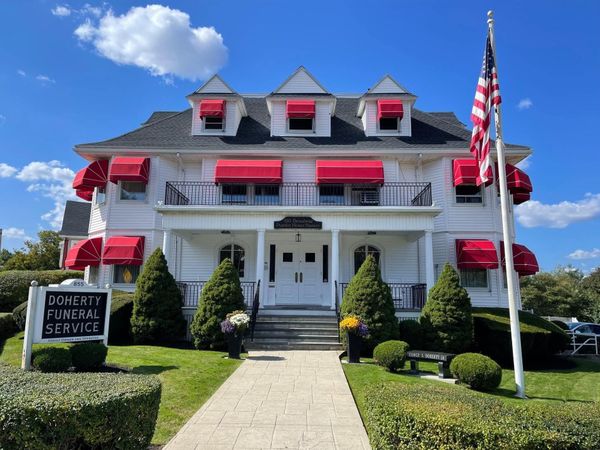 A white funeral home with red awnings and an American flag, viewed from a front walkway under a blue sky.