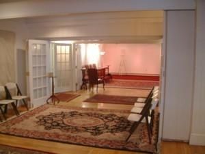 Empty room with large patterned rugs, chairs along the walls, and a doorway to a softly lit dining area